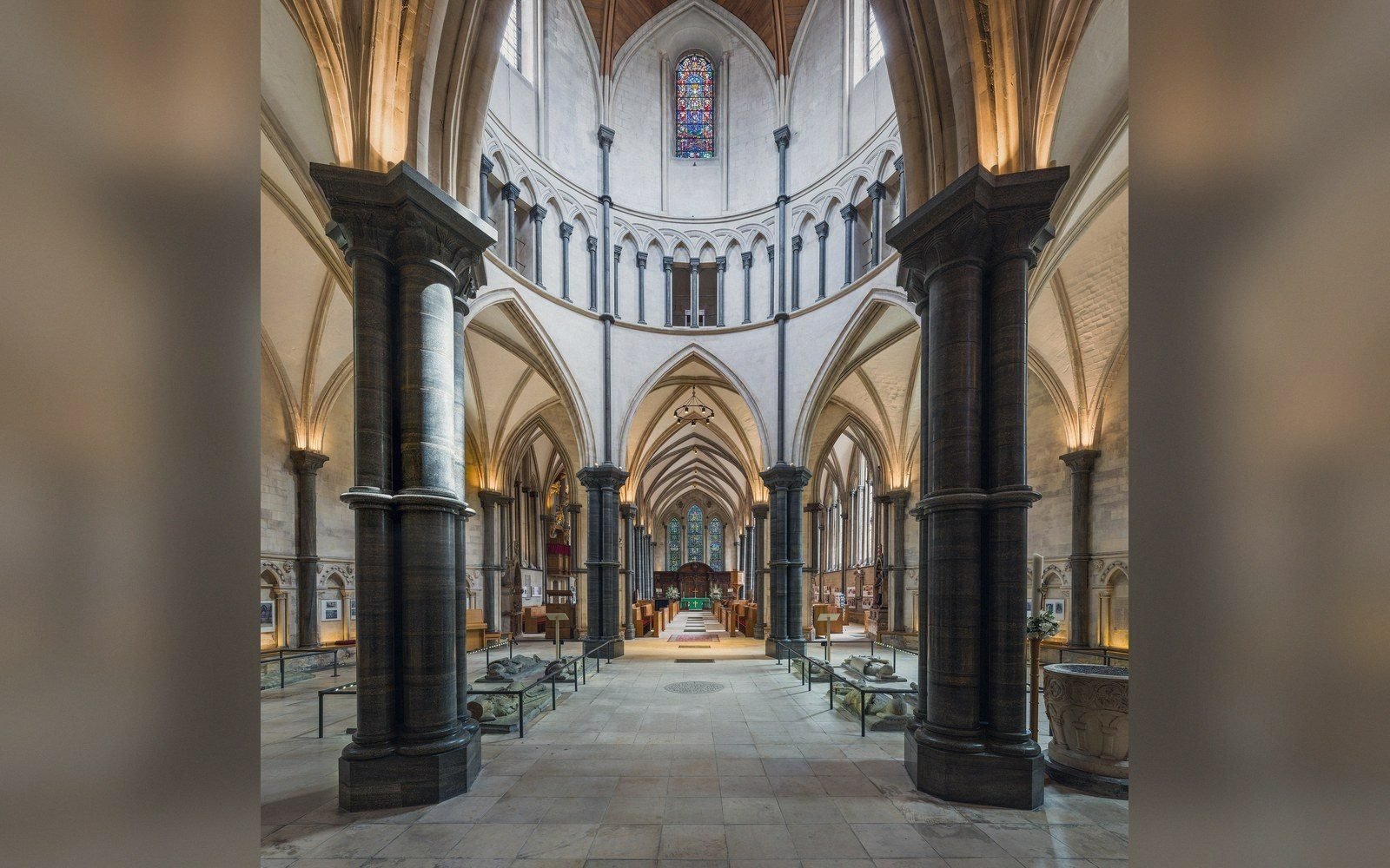 Gothic church interior with dark stone columns leading to a round apse and stained glass