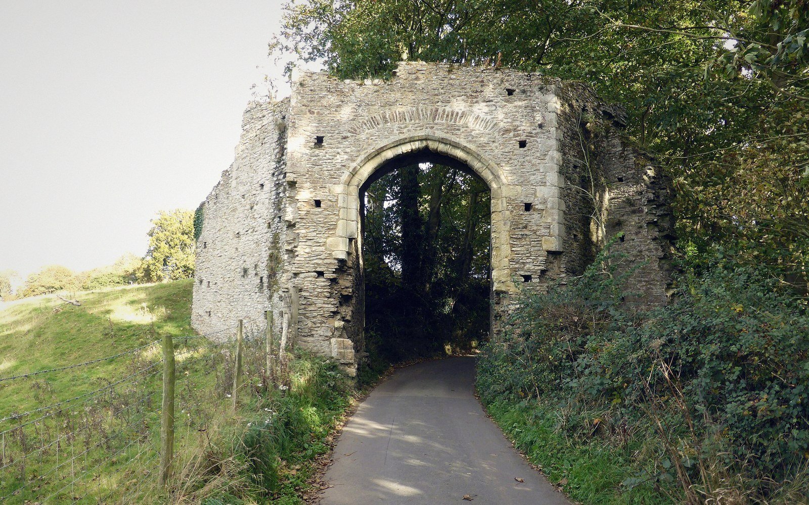 Ruined stone gateway with pointed arch straddling a narrow paved road through trees