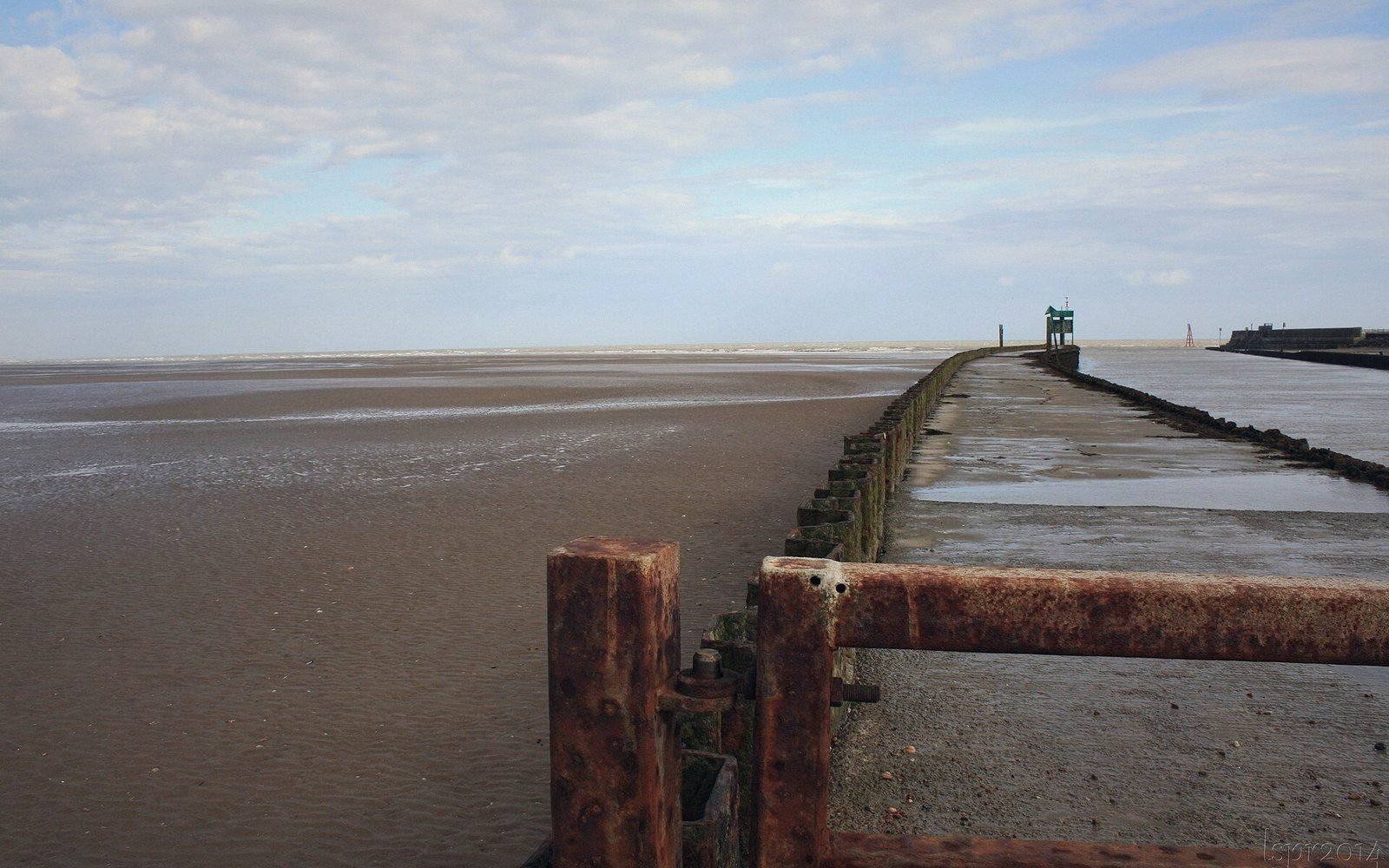 A rusted metal groyne stretches along a sandy beach toward the distant sea